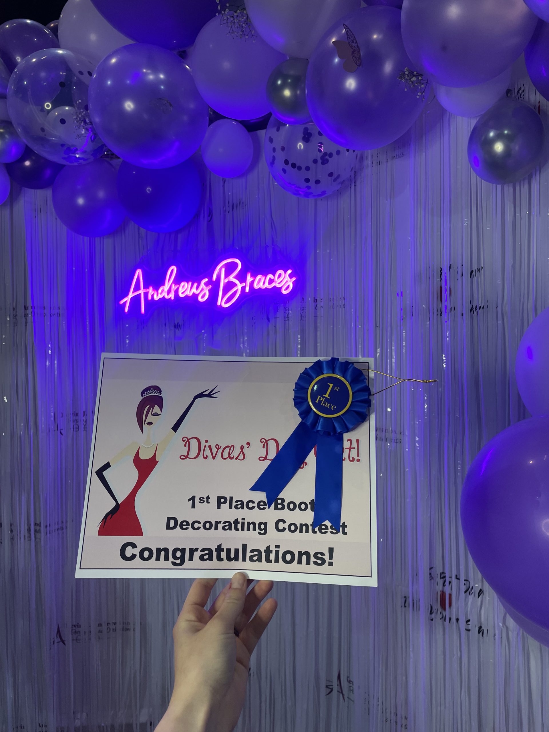 A hand is holding the 1st place award for Andrews Braces in front of a photo booth with an "Andrews Braces" neon sign